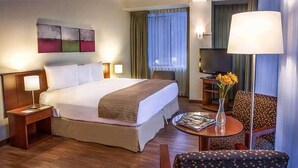 A hotel room with a large bed, bedside tables with lamps, a TV, a small table with a vase of flowers, and a chair. - Del Prado Hotel (Lima)