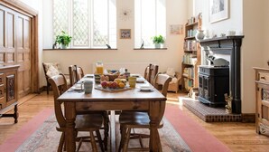 A dining room with a wooden table set for four, surrounded by chairs. There is a fireplace with a mantel, bookshelves, and a window with a view of greenery outside. - The School House (Bridgwater)