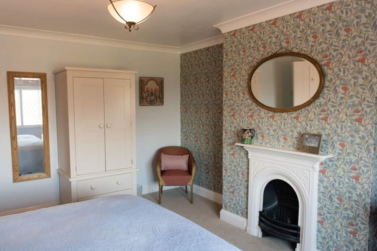 A bedroom with a bed, a white wardrobe, a chair, a fireplace, and a mirror on the wall.
