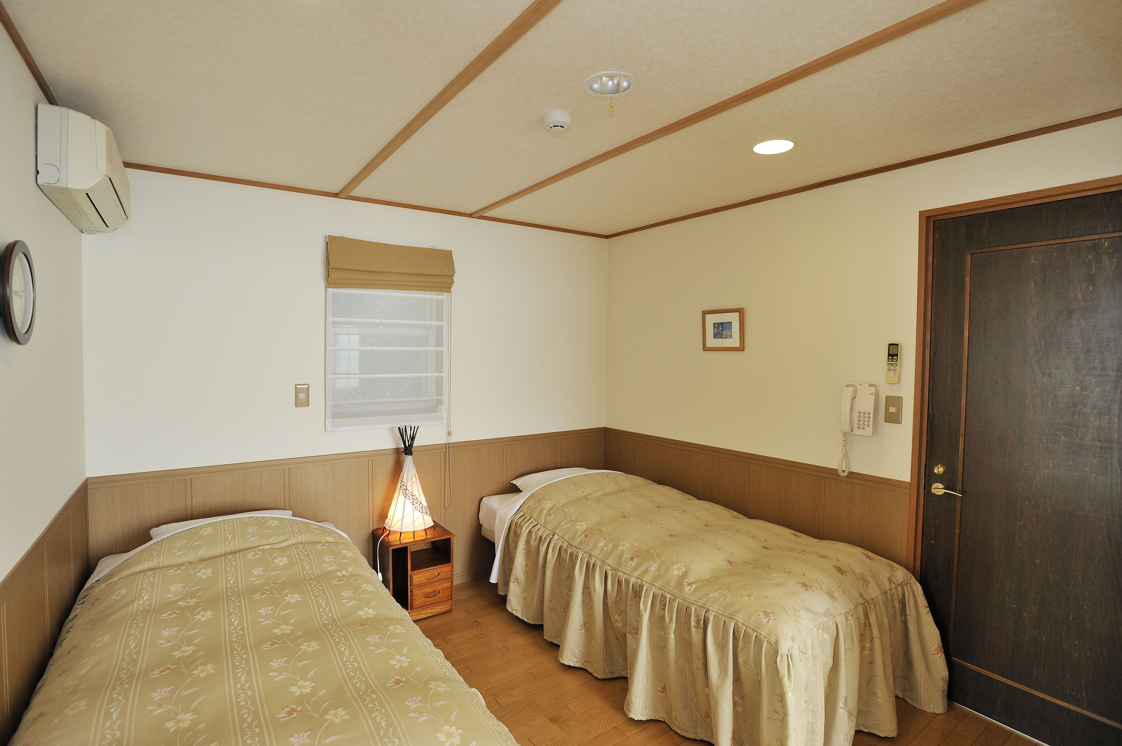 A room with two single beds, a bedside table with a lamp, a window with a blind, a wall-mounted phone, and a door.