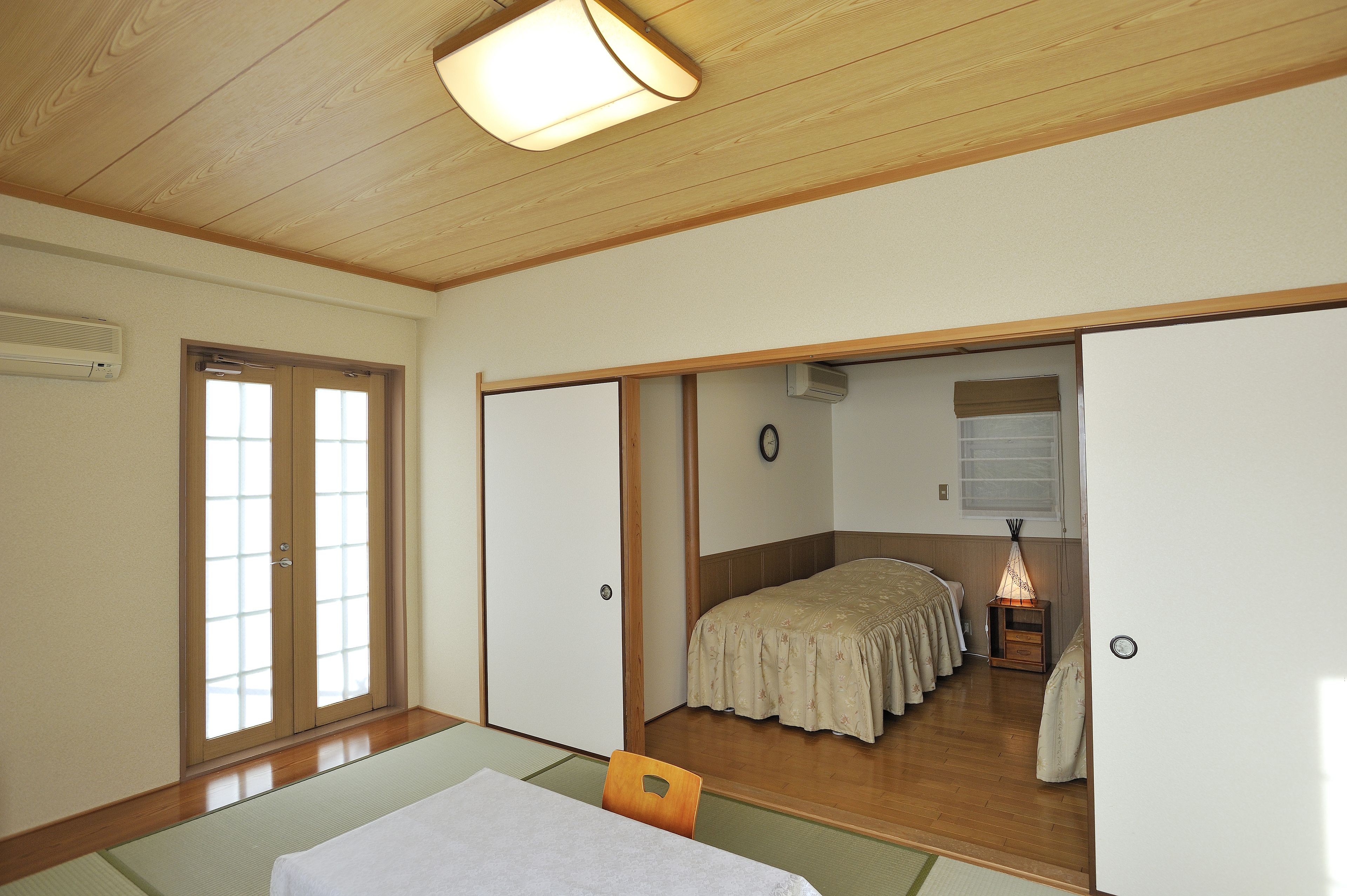 A traditional Japanese room with tatami flooring, a low table with a white tablecloth, a bed with a floral bedspread, and sliding doors.