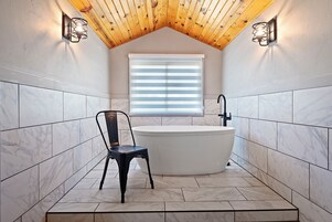 A bathroom with a freestanding bathtub, a chair, and a window with blinds.