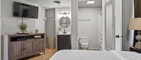 A bedroom with a bed, a TV mounted on the wall, a dresser, a mirror, and a bathroom with a toilet and shower.