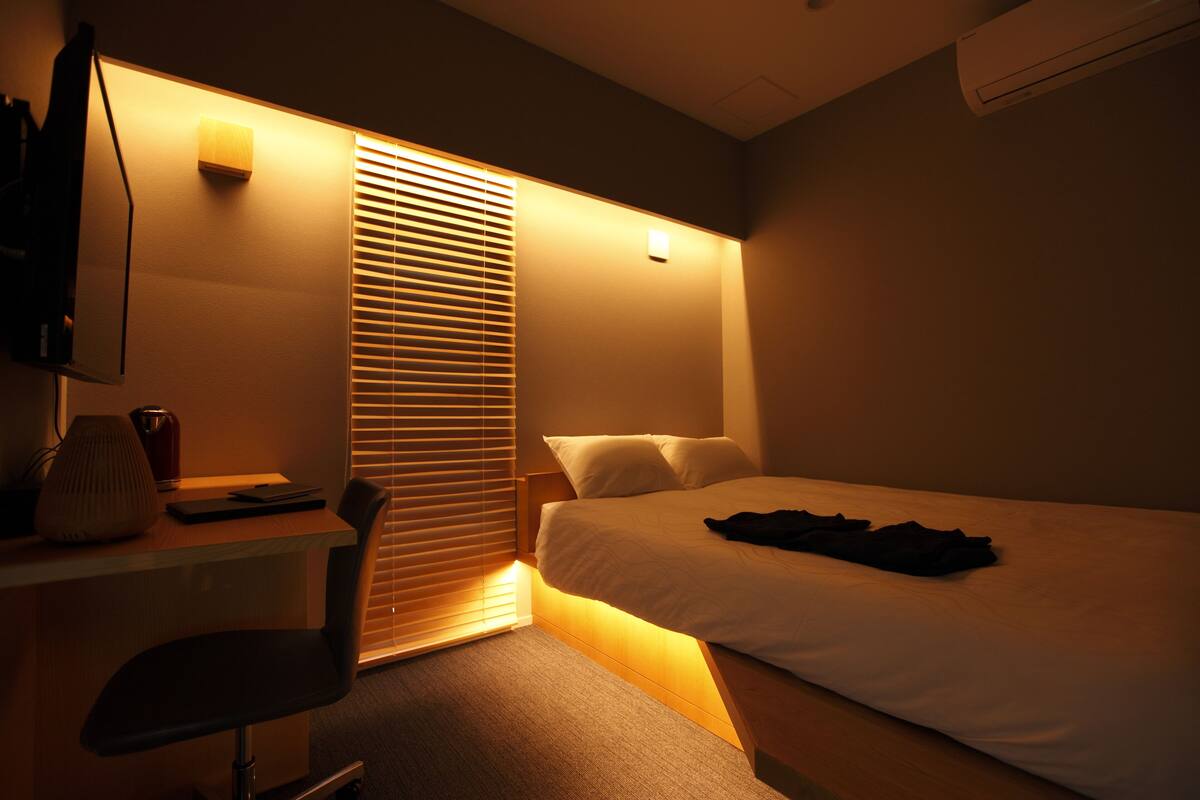 A hotel room with a bed, a desk with a chair, a lamp, and a window with blinds.