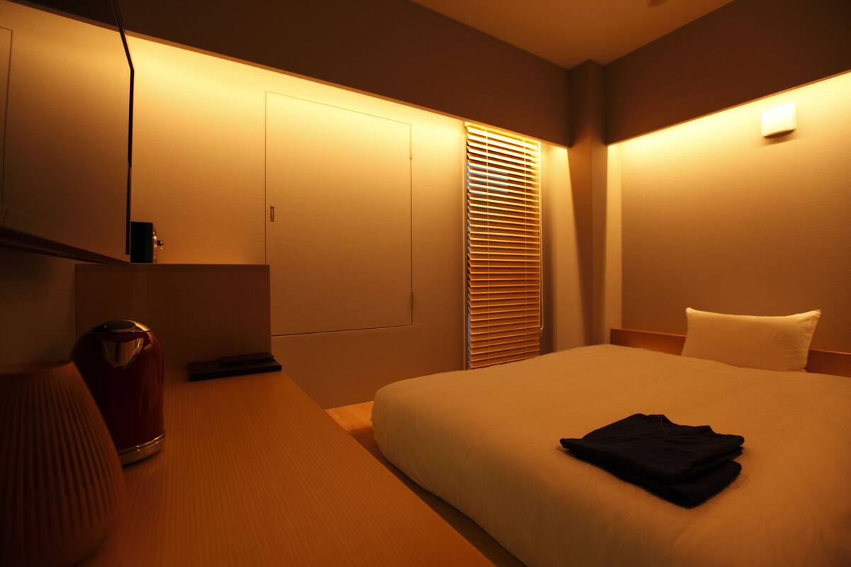 A hotel room with a bed, a nightstand, a lamp, a trash bin, and a window with blinds.