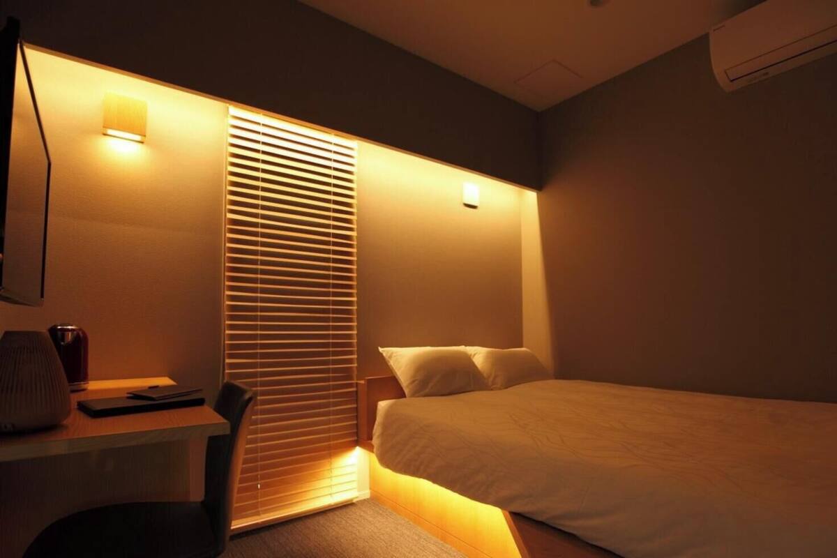 A hotel room with a bed, a desk with a lamp, a chair, and a window with blinds.