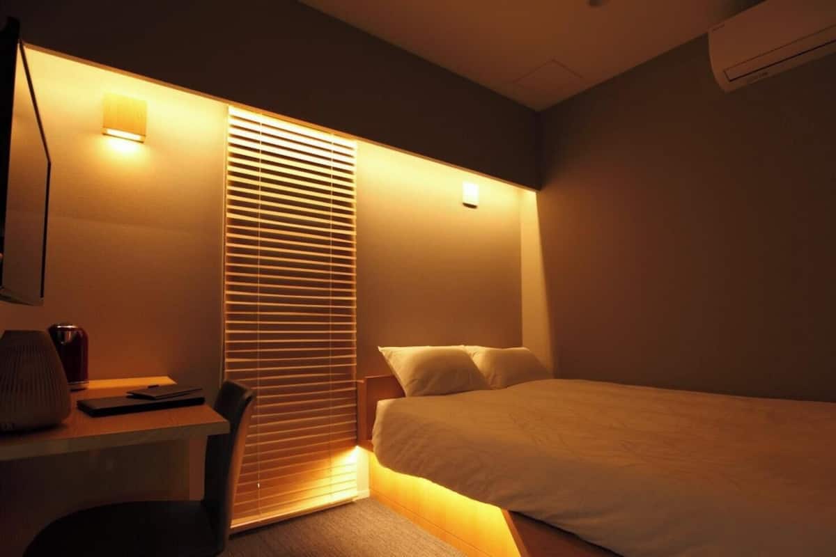 Habitación de hotel con cama, escritorio con lámpara, silla y ventana con persianas.