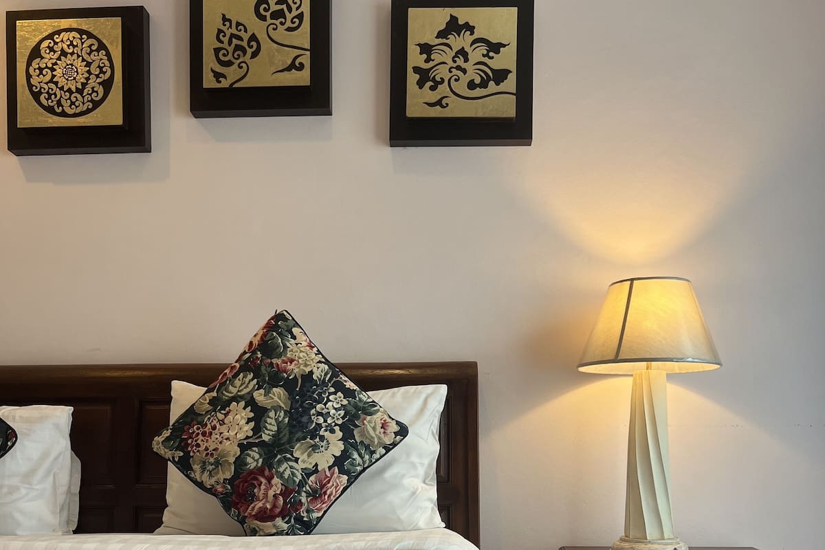 Een net opgemaakt bed met een bloemenkussen, een bedlampje en drie ingelijste kunstwerken aan de muur.