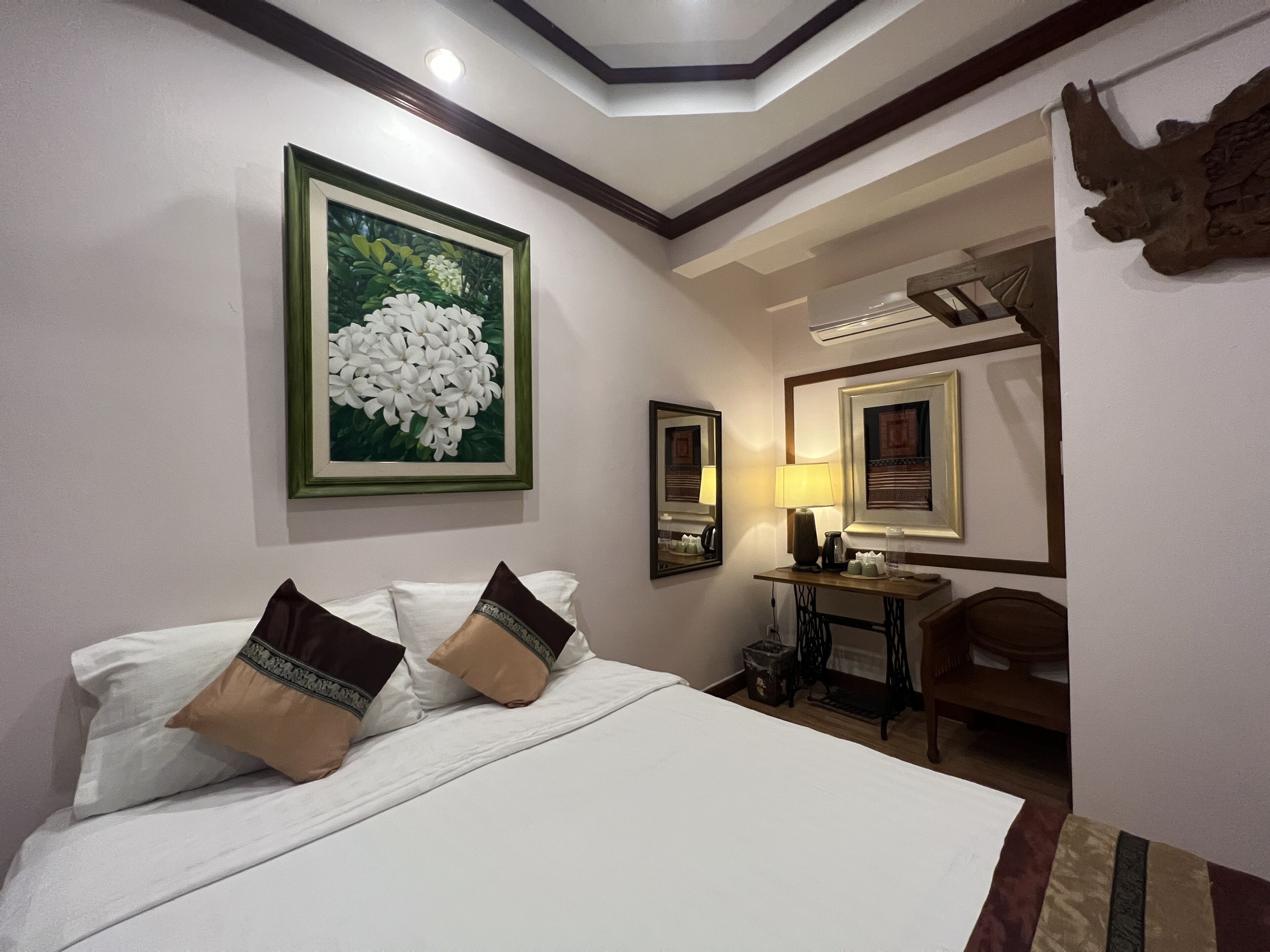 Una habitación de hotel con cama, almohadas, un escritorio con lámpara, un espejo y un cuadro en la pared.