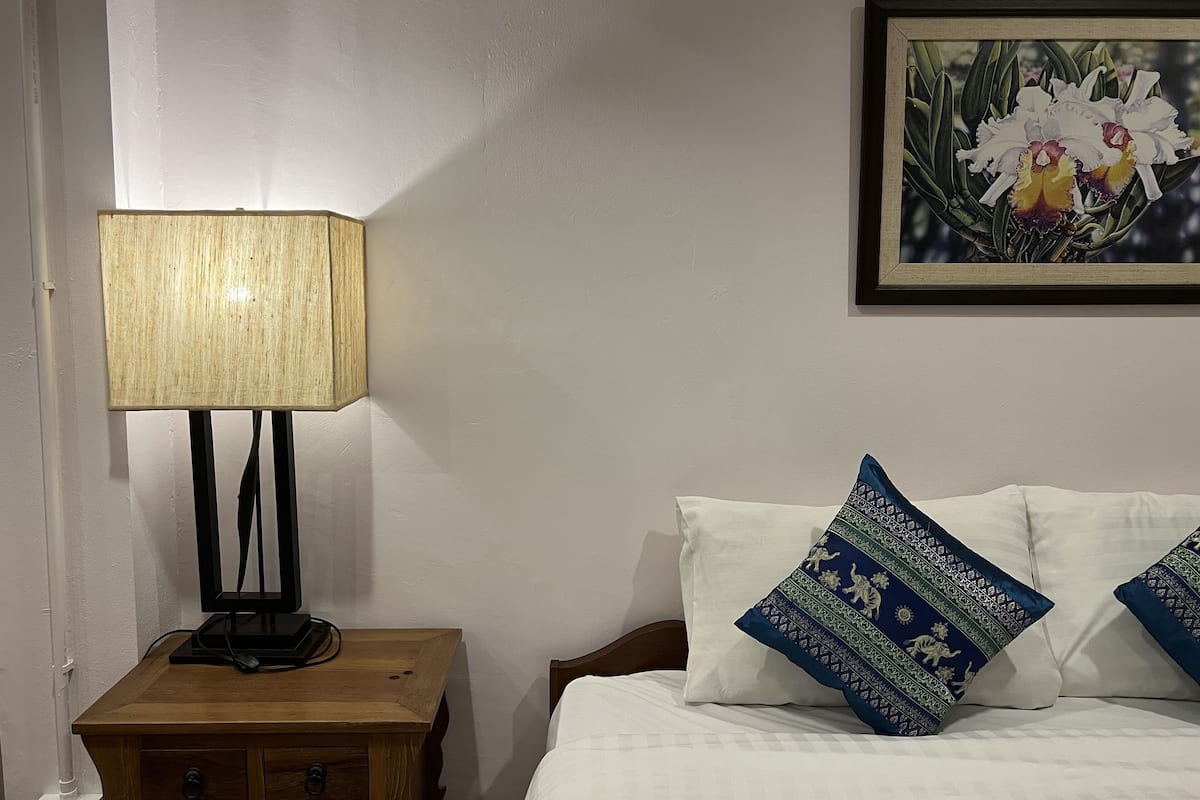 Een net opgemaakt bed met witte lakens en decoratieve kussens, een houten nachtkastje met een lamp, en een lijst met bloemkunstwerk aan de muur.