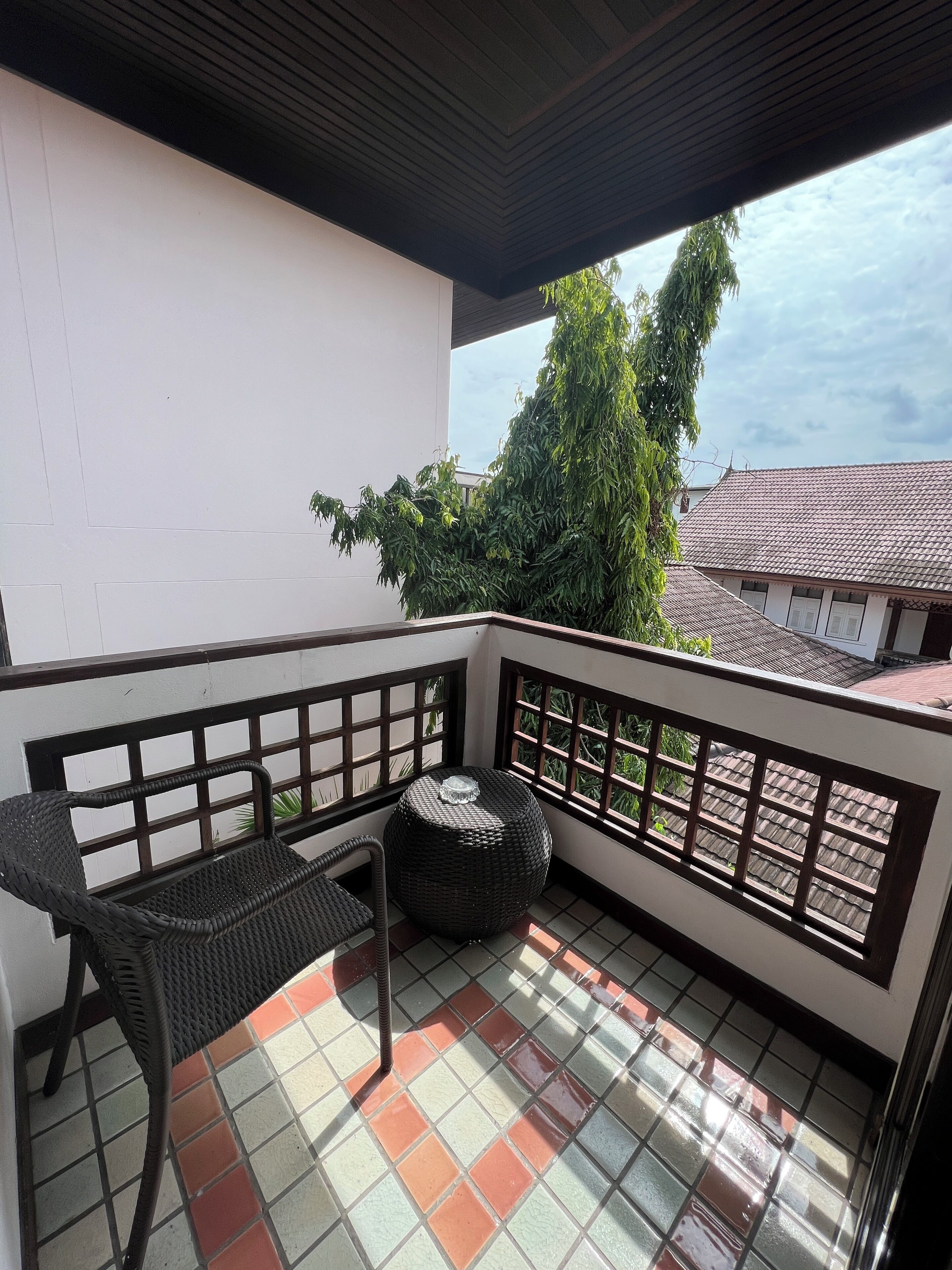 Un balcón con una silla de mimbre y una mesa redonda, piso de baldosas y vistas a edificios y árboles.