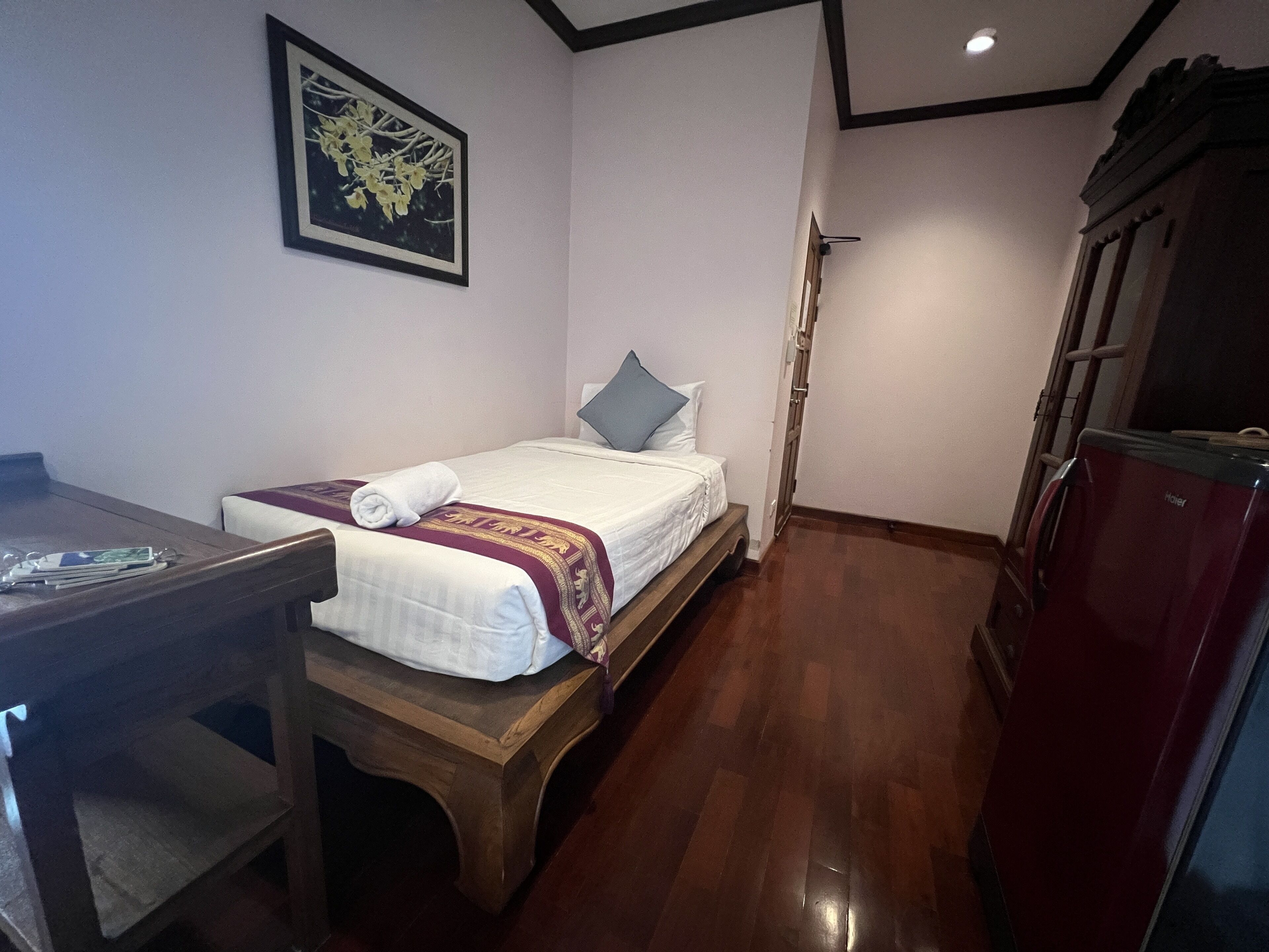 A small, neatly arranged room with a bed, a desk, a refrigerator, and a framed picture on the wall.