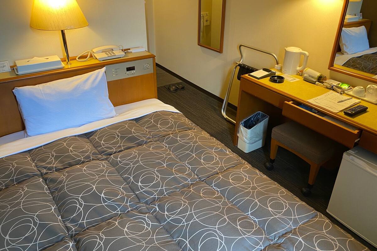 Ein Hotelzimmer mit Bett, Nachttisch, Lampe, Schreibtisch, Stuhl und kleinem Kühlschrank.