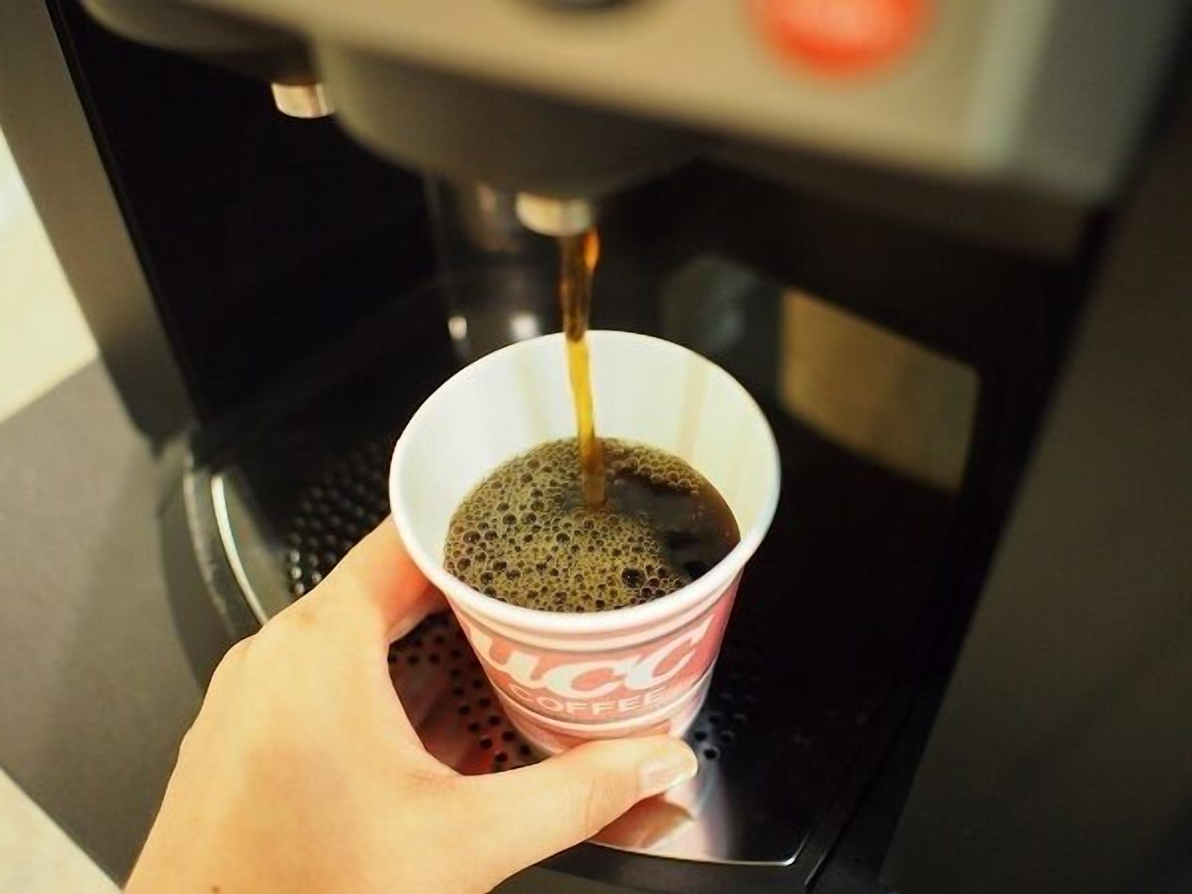 A hand holding a disposable coffee cup with a logo, positioned under a coffee machine dispensing liquid into the cup.