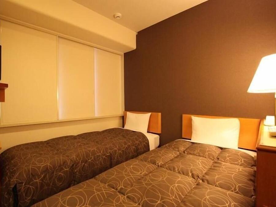 Quarto de hotel com duas camas, uma janela com persianas, mesa de cabeceira com abajur e televisão em uma estante.