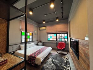 Une chambre d’hôtel moderne équipée d’un lit, d’un fauteuil suspendu, d’une télévision et d’une fenêtre aux vitraux colorés.