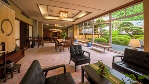 Un vestíbulo de hotel con una decoración interior tradicional japonesa, que presenta esteras de tatami, un mostrador de recepción y vistas a un jardín exuberante a través de grandes ventanales.