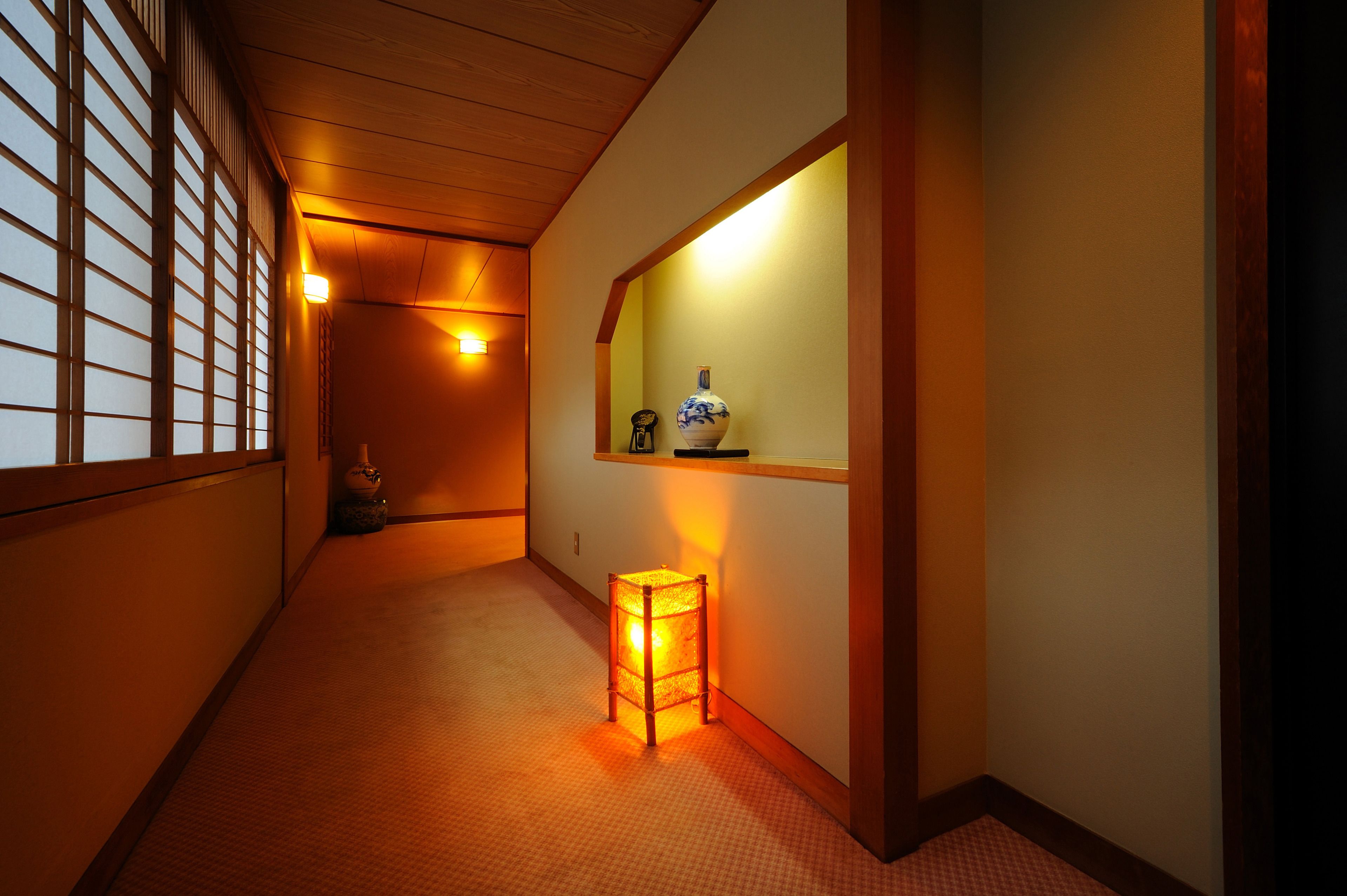 A narrow hallway with tatami flooring, a small illuminated lantern, and a shelf with a vase and a remote control.