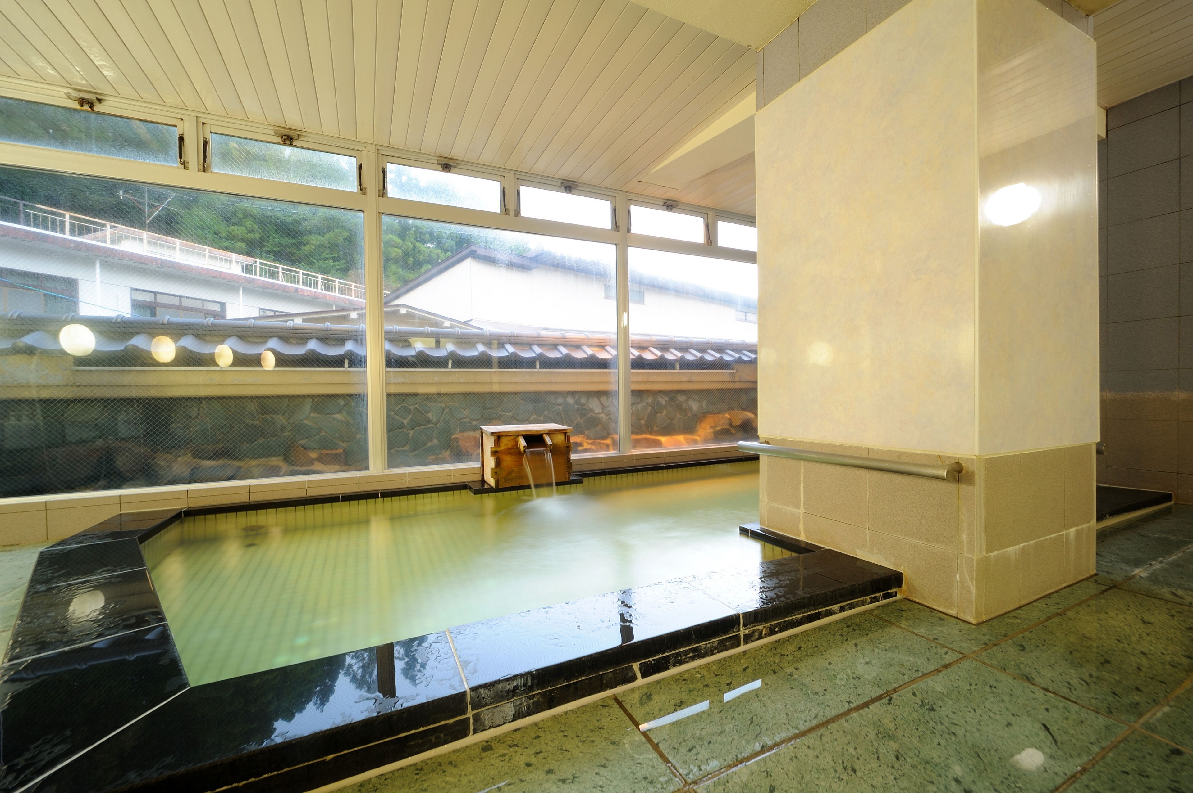 A traditional Japanese onsen with a stone bath, wooden benches, and a view of a traditional building through large windows.