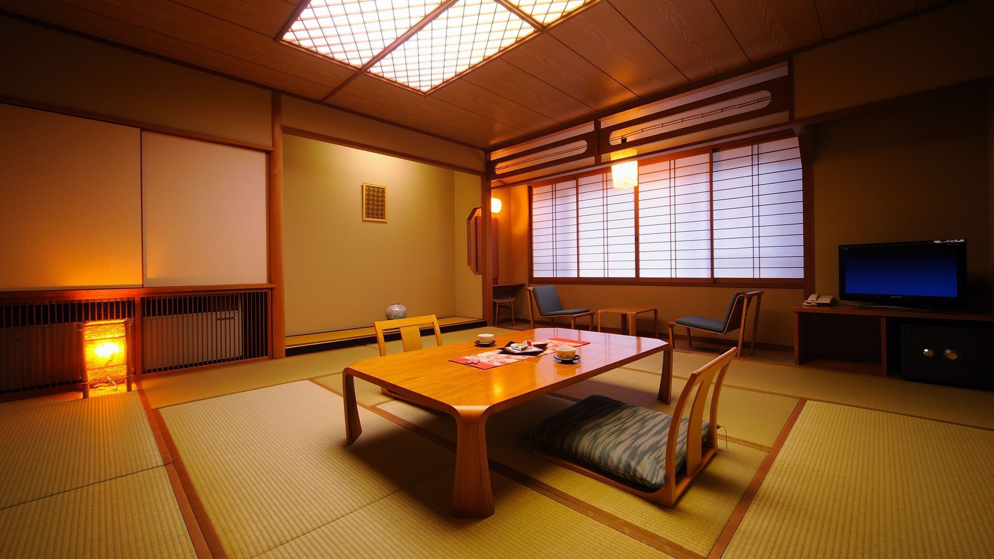 A traditional Japanese room with tatami flooring, a low table, chairs, a television, and sliding doors.