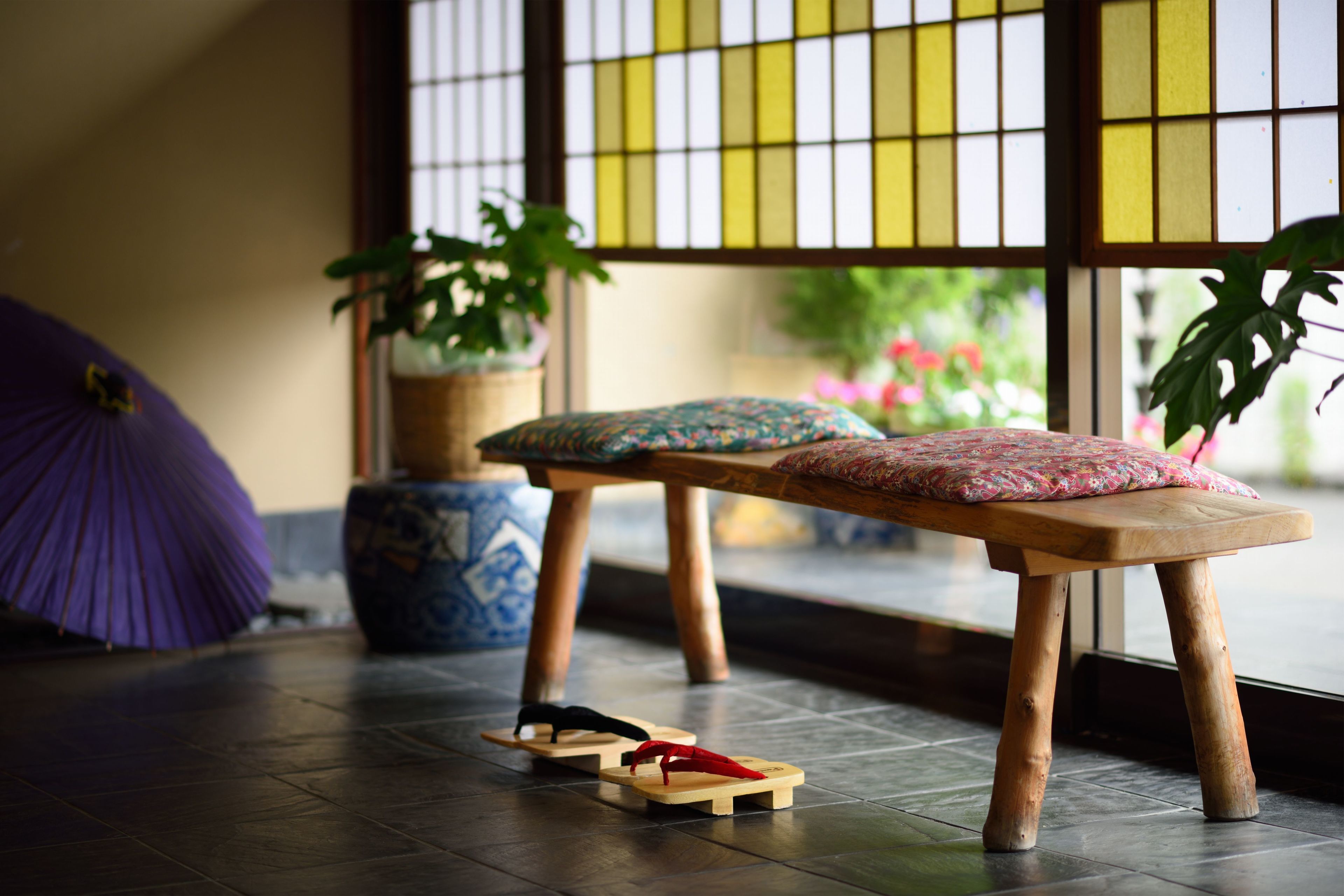 A wooden bench with cushions, a purple umbrella, and potted plants in a room with a tiled floor and a window with a grid pattern.