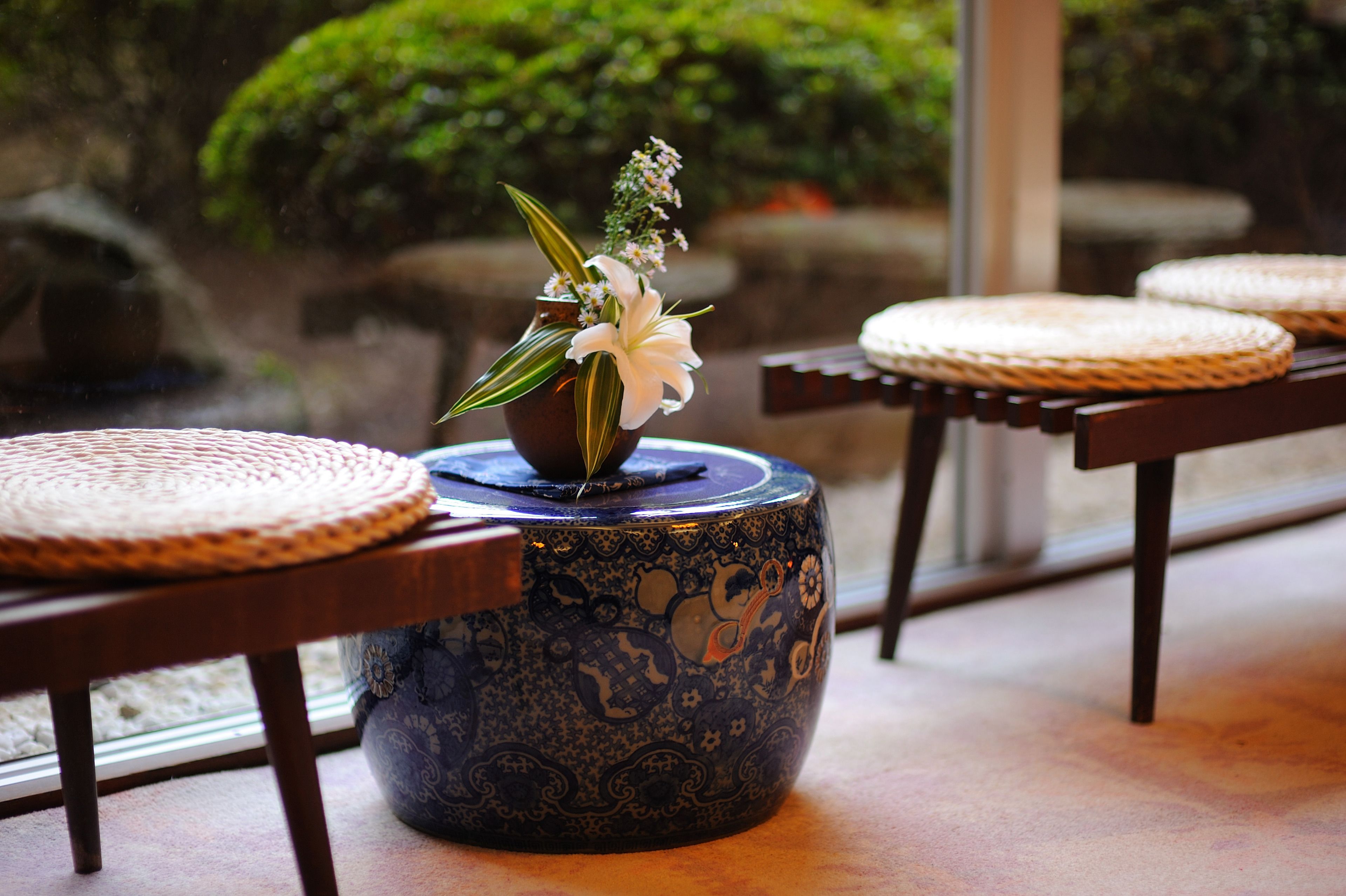 Una tradizionale stanza in stile giapponese con tatami, un vaso ceramico blu con un’esposizione floreale e vista su un giardino attraverso una porta vetrata.