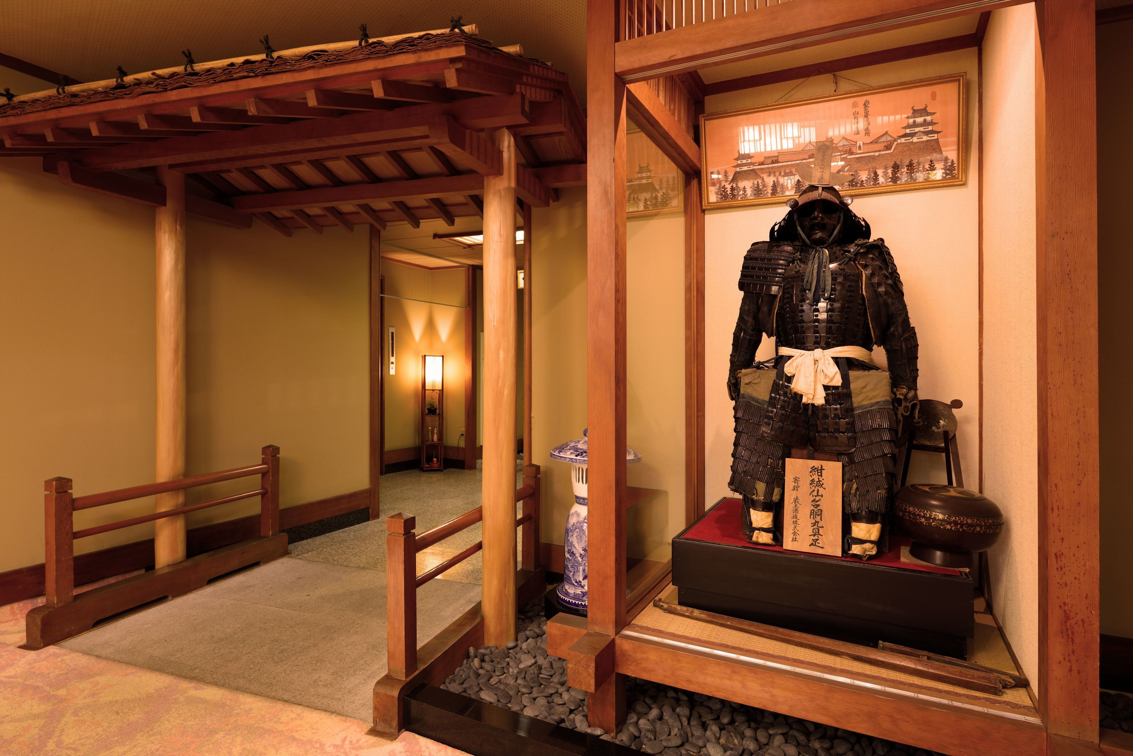 Una habitación tradicional japonesa con suelo de tatami, techo con vigas de madera y una vitrina que exhibe una armadura de samurai y un rollo.