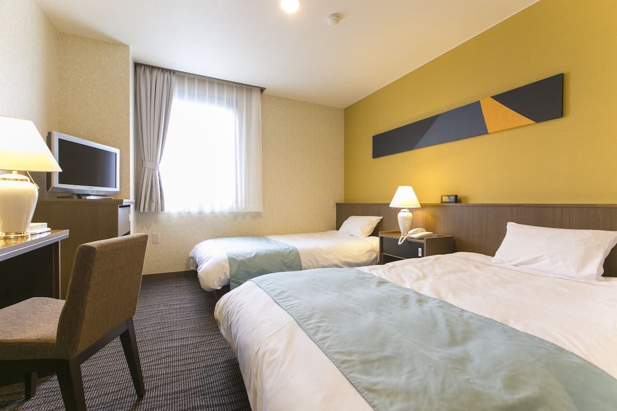 A hotel room with two beds, a desk with a TV, a chair, a lamp, and a wall-mounted phone.