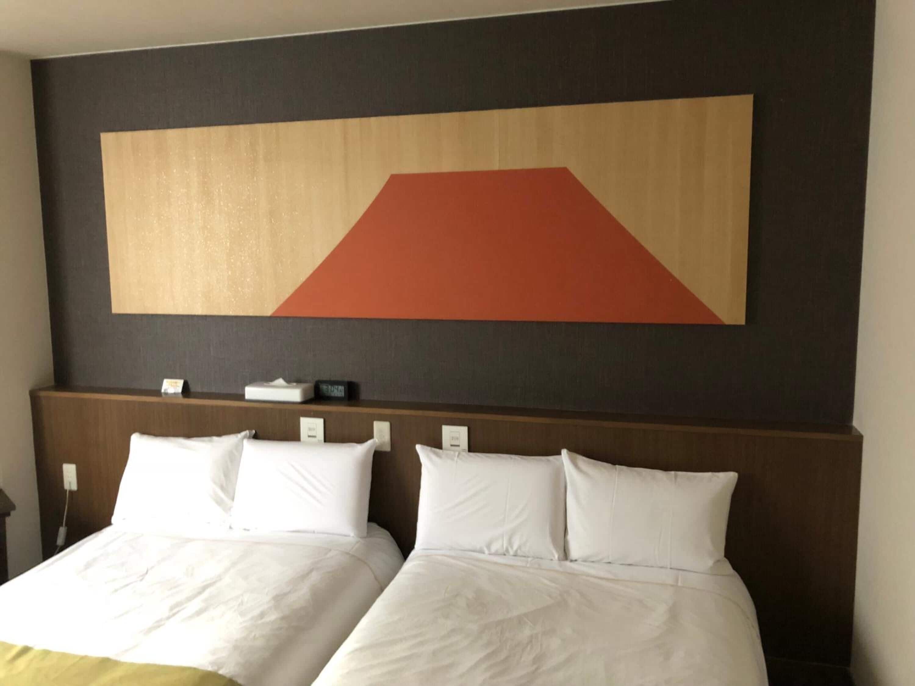 Quarto de hotel com duas camas, um quadro abstrato grande na parede e uma mesa de cabeceira com um relógio e uma caixa de lenços.