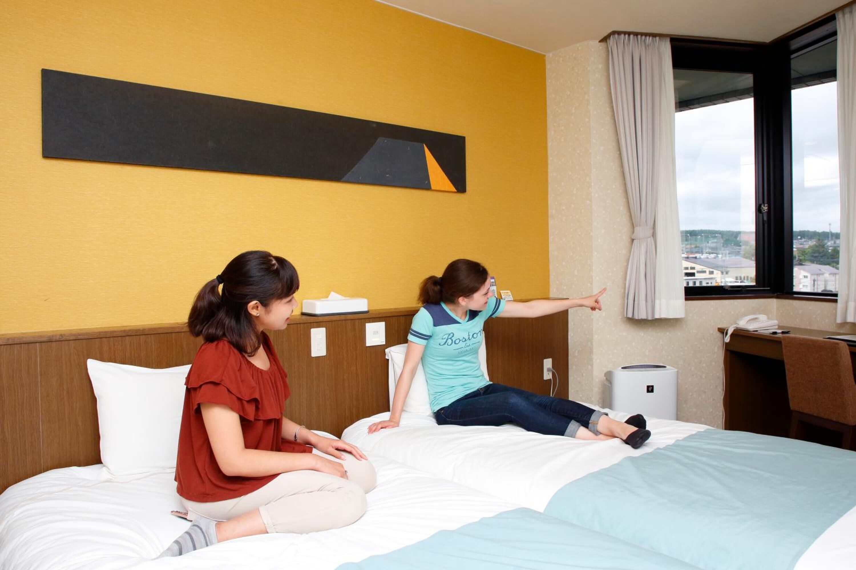 A hotel room with a bed, a desk, a chair, a window with curtains, and a wall-mounted artwork.