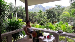 A person sitting on a white plastic chair under a shaded area, reading a book. There is a small white table with a hat and a bowl on it. The background features lush greenery and a mountainous landscape. - Aonang Cliff Mountain New Resort (Krabi)