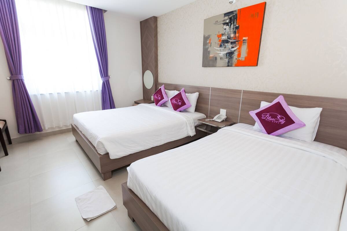 A hotel room with two beds, a nightstand with a phone, a window with purple curtains, and a painting on the wall.