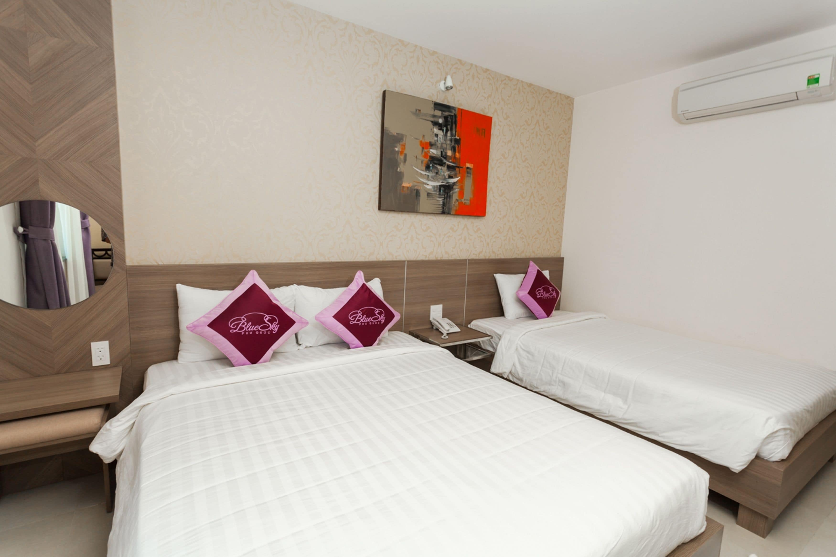 A hotel room with two beds, each with white bedding and pink pillows, a mirror, a painting on the wall, and an air conditioning unit.