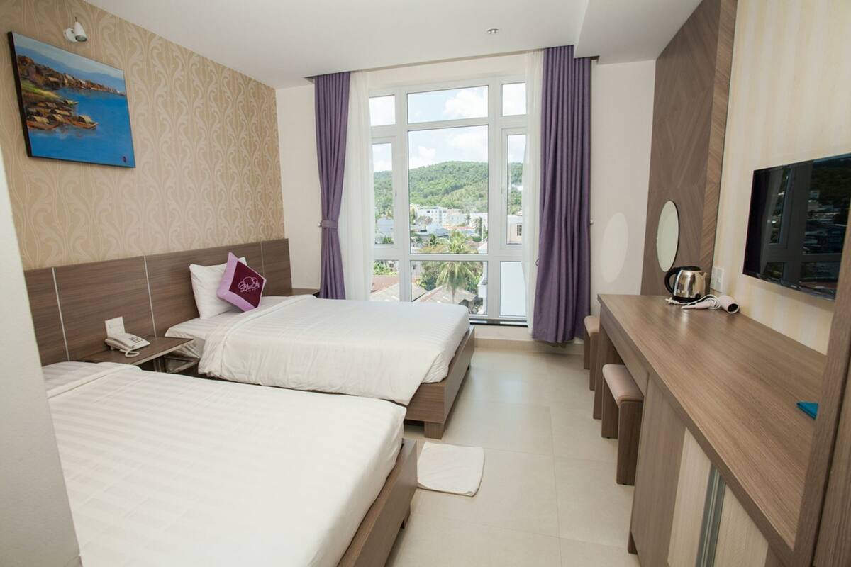A hotel room with two beds, a desk, a TV, and a window with a view of a green landscape.