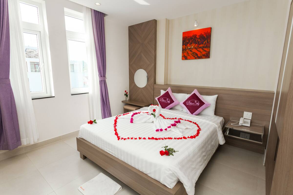 A neatly made bed with a heart-shaped flower arrangement, a bedside phone, and a wall-mounted painting.