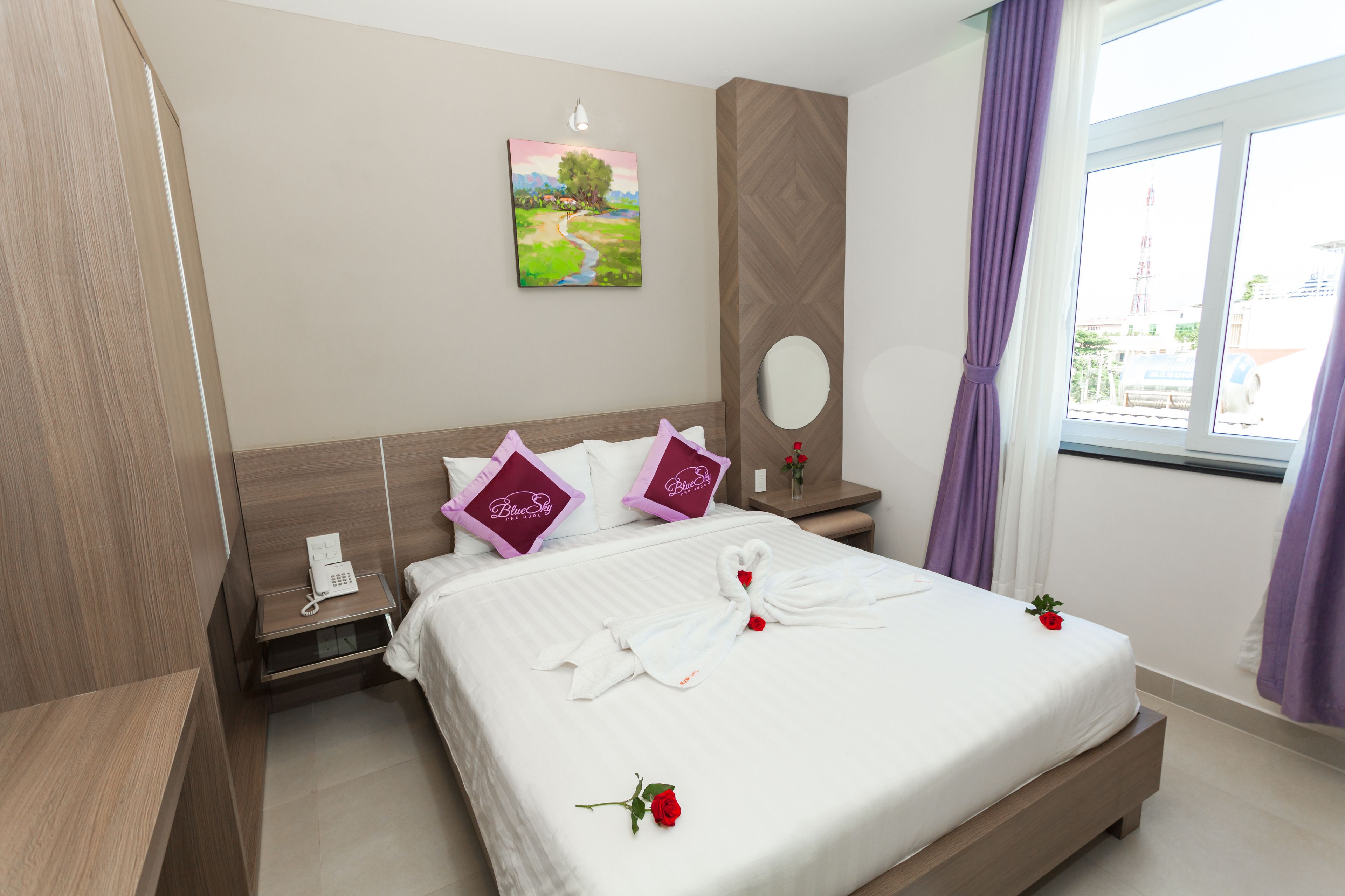 A hotel room with a neatly made bed, a bedside table with a phone, a wall-mounted mirror, and a window with curtains.