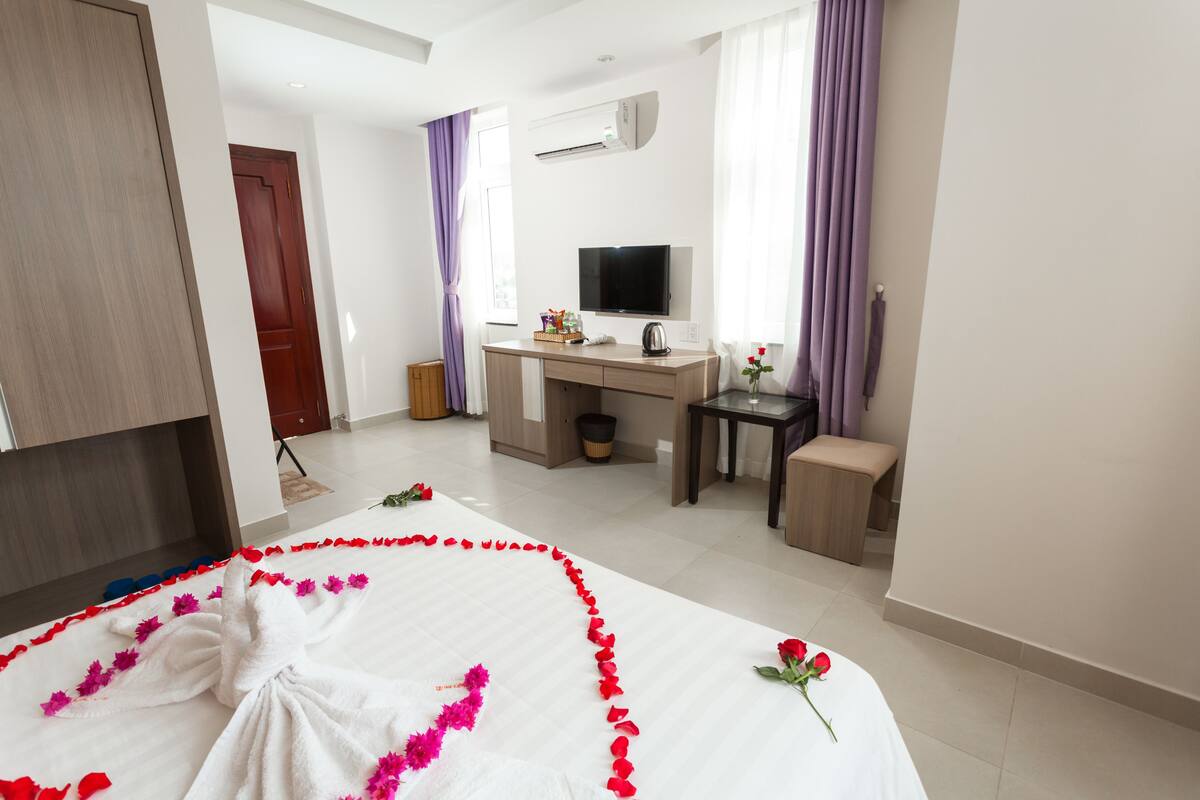 A hotel room with a neatly made bed, a desk with a computer, a TV, and a small table with a vase of flowers.