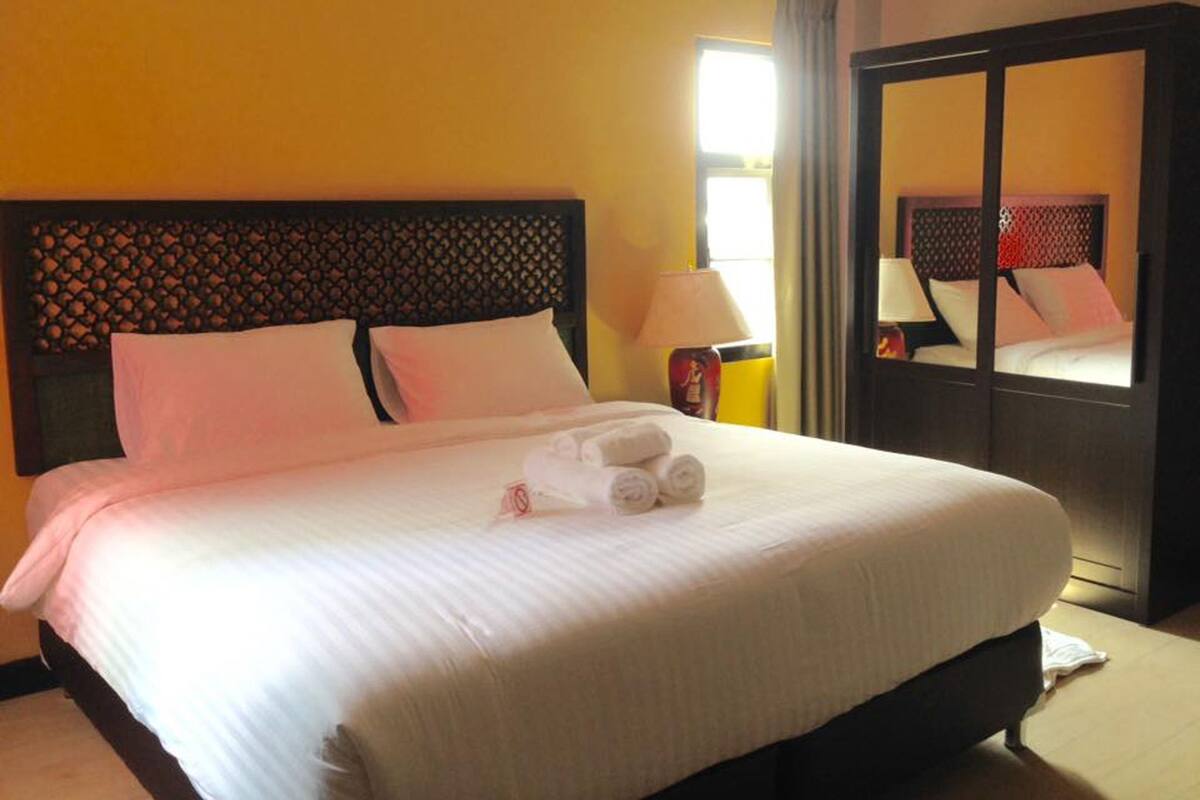 Una cama bien hecha con sábanas blancas y toallas enrolladas, un cabecero de madera con un diseño geométrico, una lámpara de noche y un espejo grande en la pared.