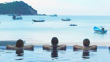 Una piscina con tres personas relajándose, con vistas al mar tranquilo y varios barcos.