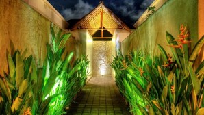 A corridor with a tiled floor, flanked by green plants and a decorative wall with a carved design. - Villa Lestari (Kerobokan)