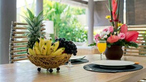 A dining table with a wicker fruit basket containing bananas, grapes, and a pineapple. There is a glass of orange juice and a vase with flowers. - Villa Lestari (Kerobokan)