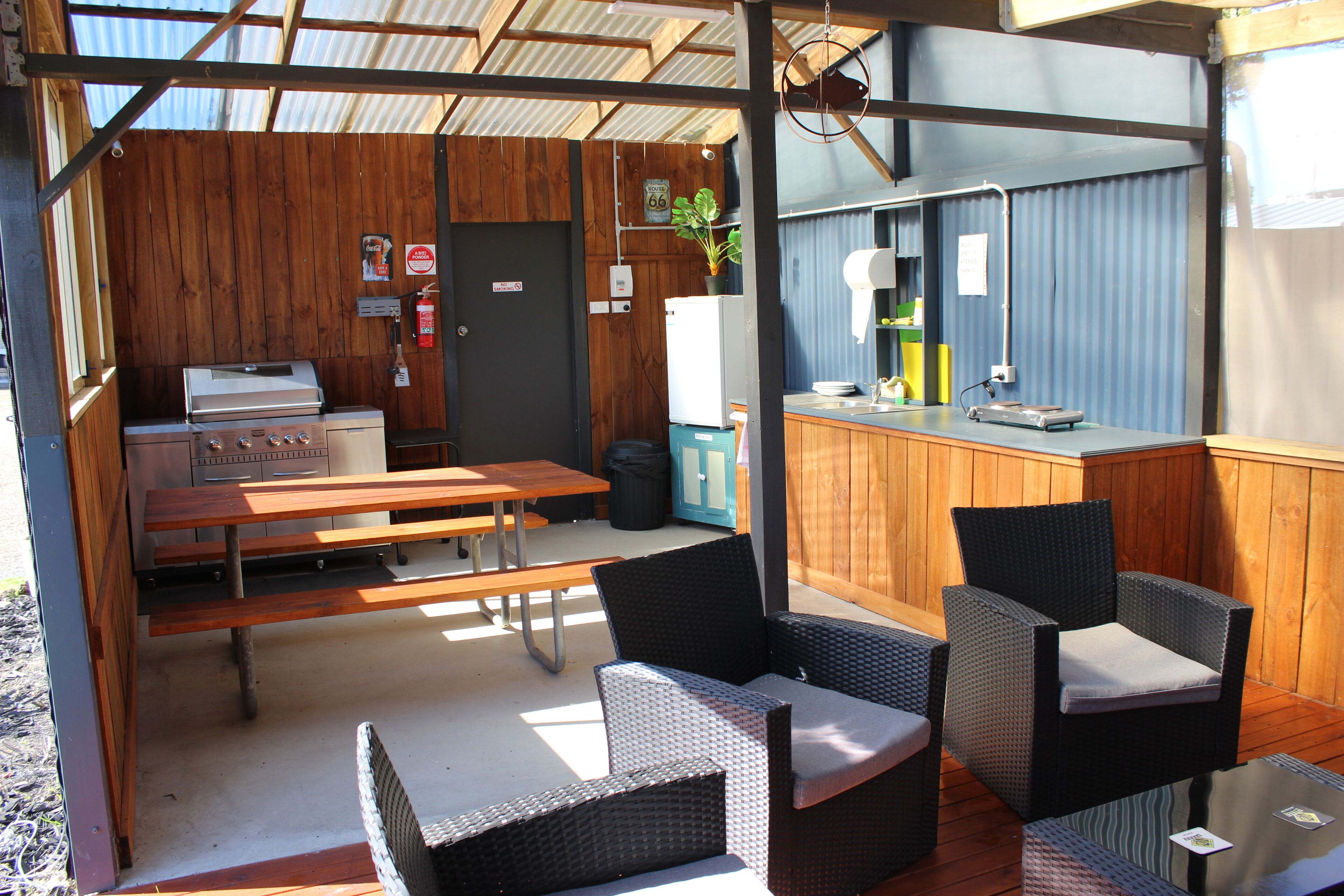 A covered outdoor area with a barbecue grill, wooden tables, wicker chairs, a bar area, and a refrigerator.