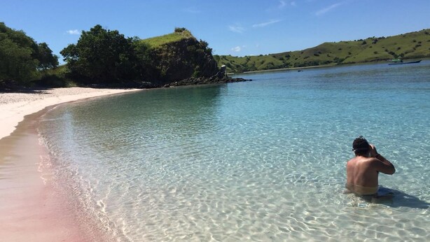 A beach with clear water, a person in the water, and a hilly landscape in the background. - Komodo Cruise Boat (Labuan Bajo)