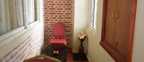 Un estrecho pasillo con pared de ladrillo, una puerta de madera, una silla roja y una pequeña mesa con un jarrón de flores.