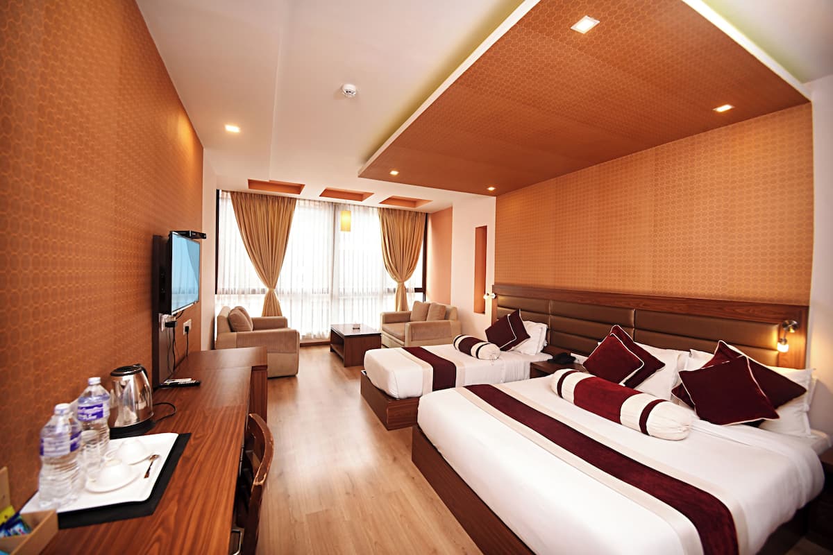 A hotel room with a wooden floor, a large bed with red and white bedding, a sofa, a TV, and a desk with a kettle and water bottles.