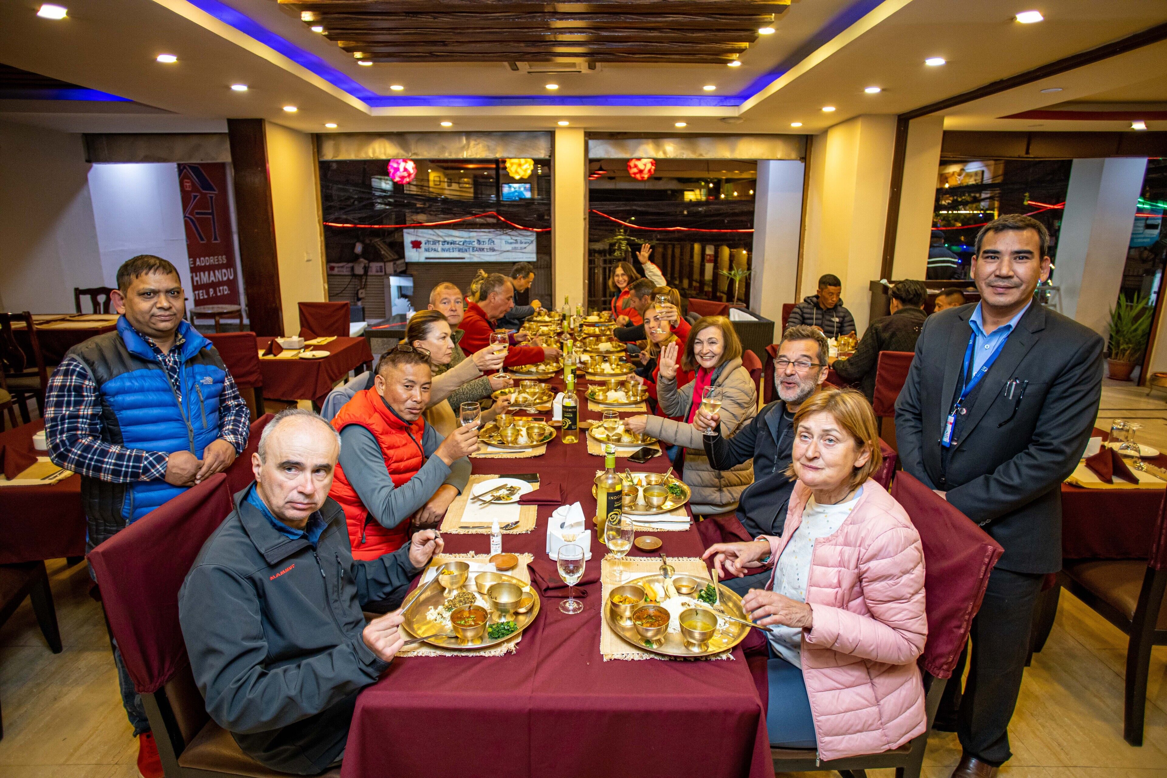 A group of people seated around a table with traditional dishes, in a well-lit indoor dining area with a modern ceiling design.