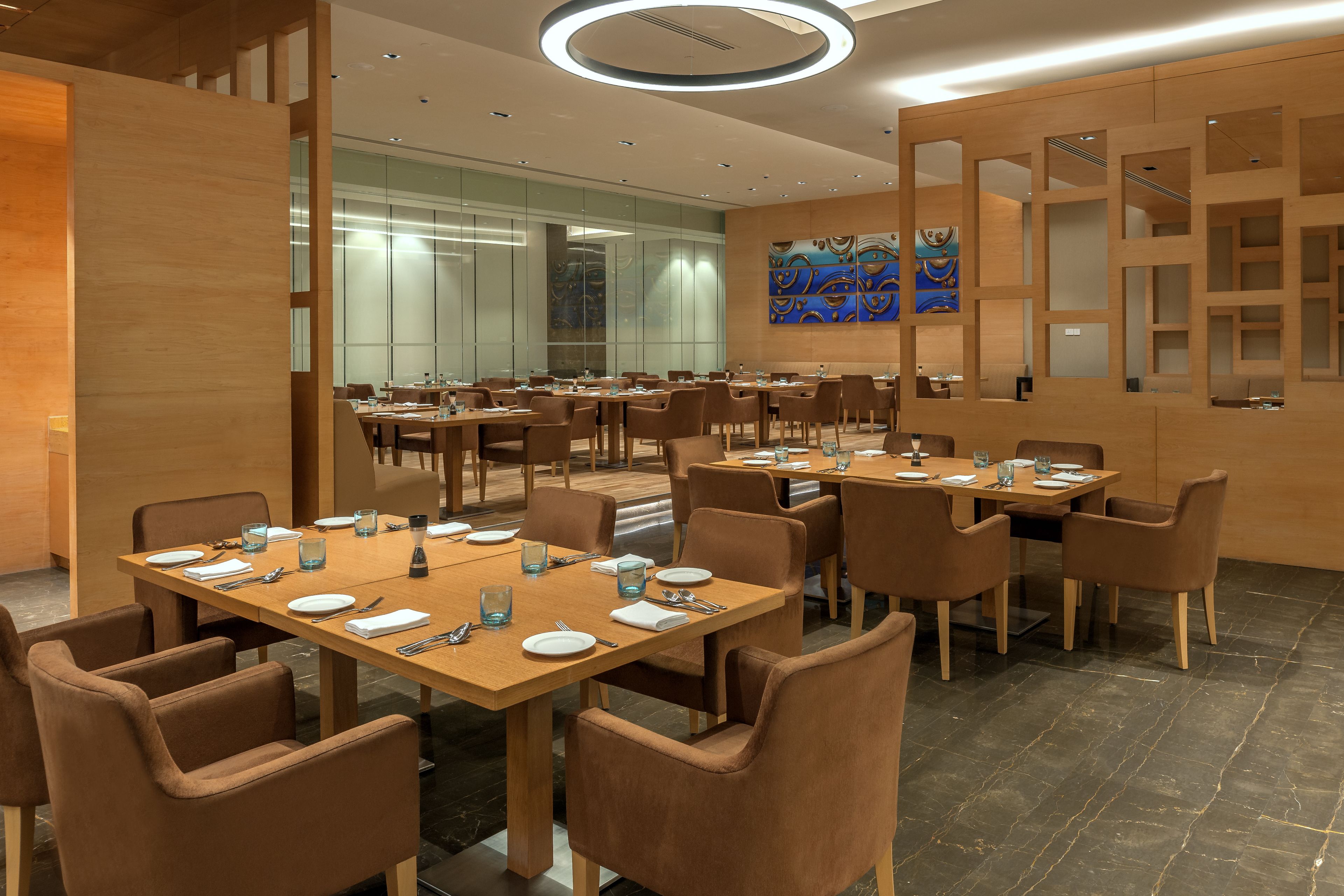 A modern hotel dining area with multiple tables and chairs, glass partitions, and a contemporary art piece on the wall.