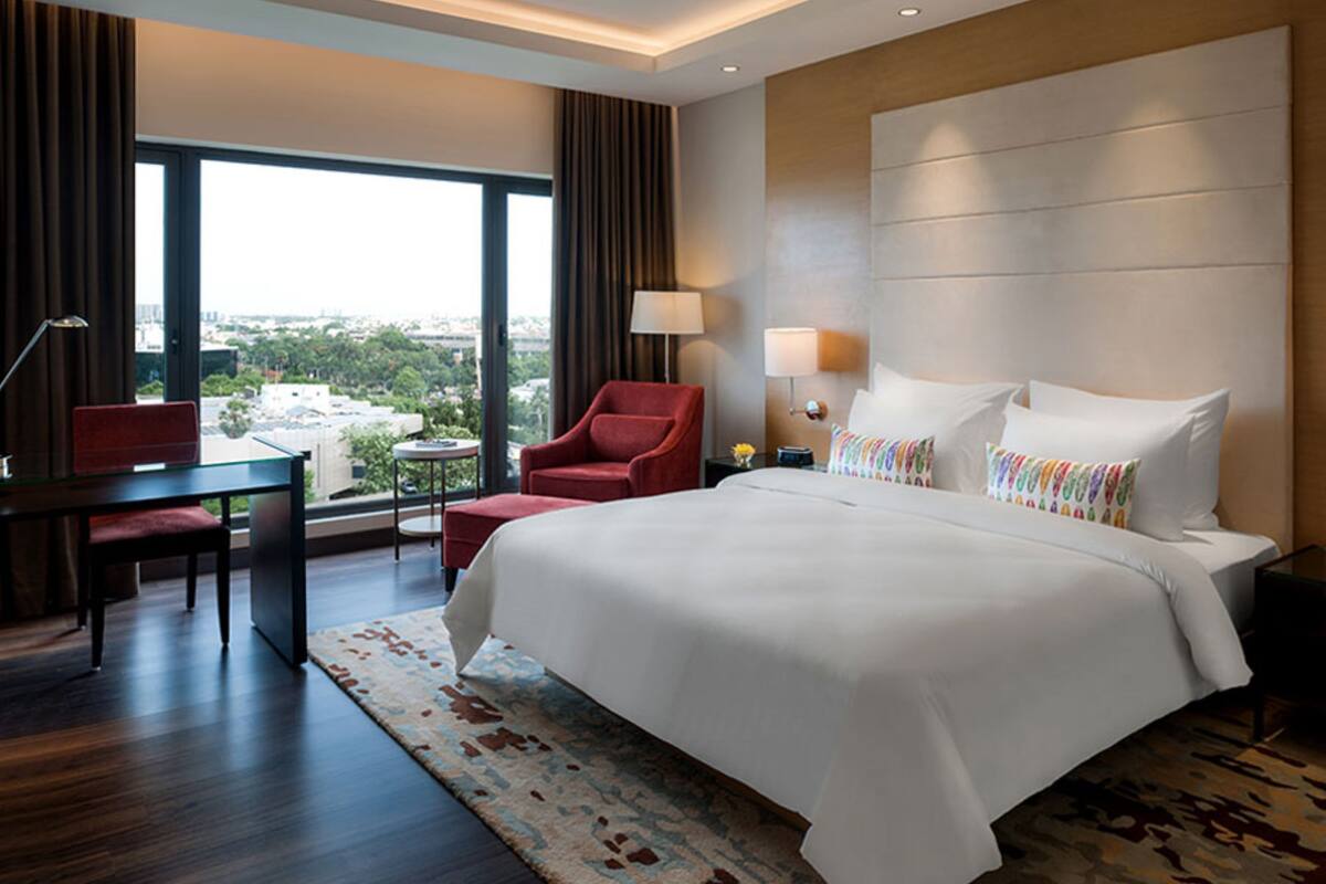 A hotel room with a large bed, a desk, a chair, and a view of the city through large windows.