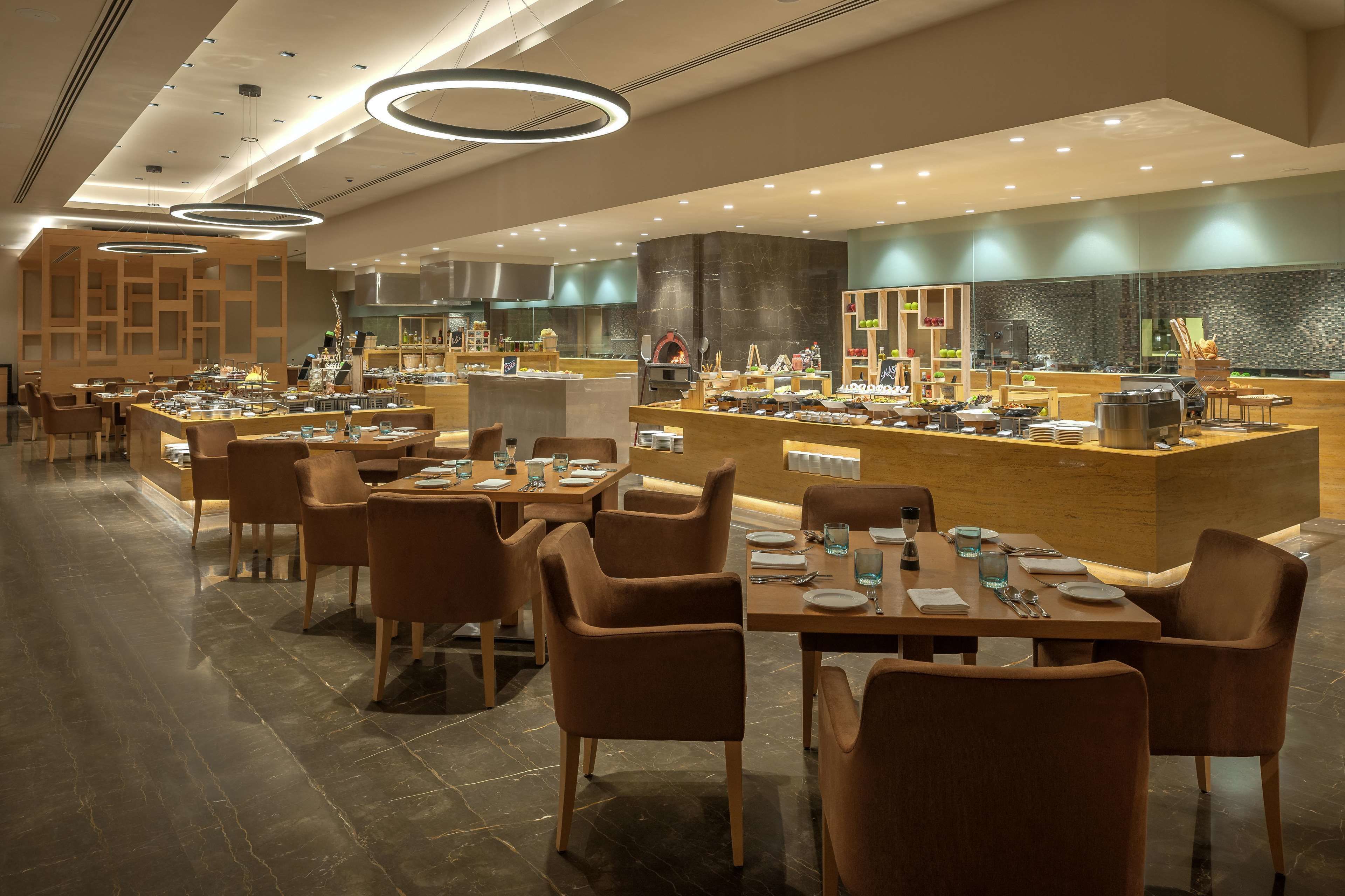 A modern restaurant with multiple dining tables and chairs, a central counter with food displays, and a kitchen area in the background.