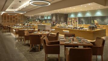 A modern restaurant with multiple dining tables and chairs, a central counter with food displays, and a kitchen area in the background.